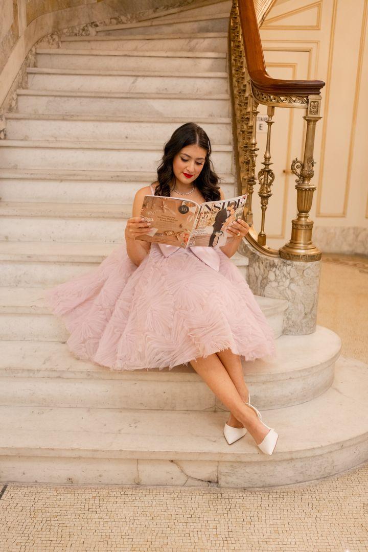 Jesse wearing a pink dress reading a magazine on a staircase.